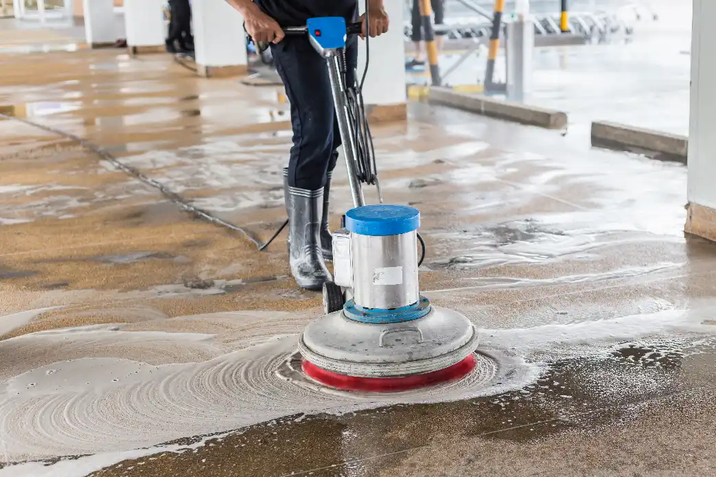 A worker in tall rubber boots operates a rotary floor scrubbing machine with a red pad, spreading soapy cleaning solution across a concrete floor in a covered parking or industrial area.